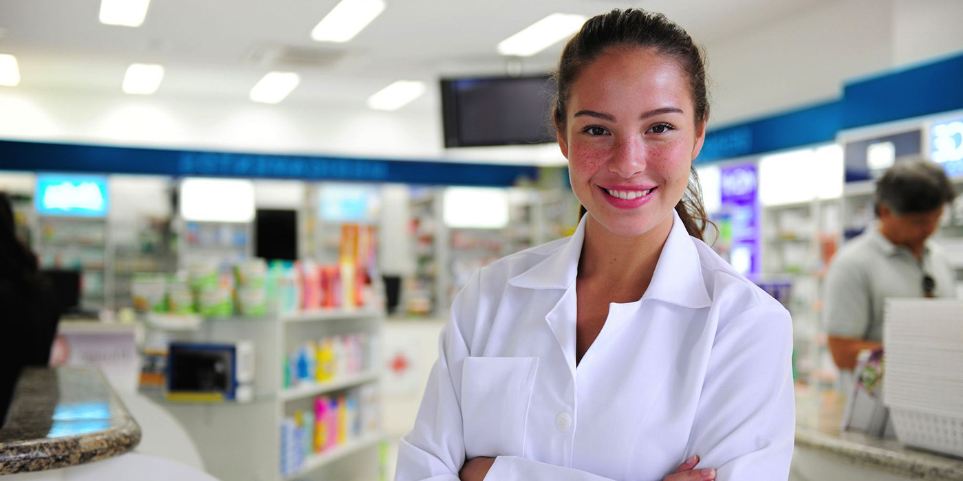 A pharmacist standing at the desk of a pharmacy smiling at the camera